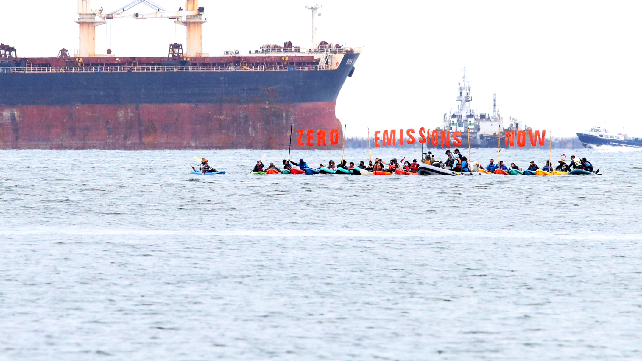 A landscape of ocean on an overcast day in Southern California. 20+ colorful kayaks are in the distance and riders hold a large orange sign reading "Zero Emissions Now." There is a large oil tanker and other marine vessels in the background.
